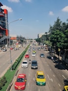 A busy urban street with multiple lanes of traffic, including cars and motorcycles. On each side of the road, there are buildings, trees, and pedestrians walking on sidewalks. Some vehicles are taxis, marked with distinctive paint colors. Traffic is relatively organized, and the weather appears to be clear with a blue sky and a few clouds.
