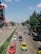 A busy urban street with multiple lanes of traffic, including cars and motorcycles. On each side of the road, there are buildings, trees, and pedestrians walking on sidewalks. Some vehicles are taxis, marked with distinctive paint colors. Traffic is relatively organized, and the weather appears to be clear with a blue sky and a few clouds.