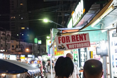 A busy nighttime street scene with a prominent 'For Rent Motorbike' sign hanging in the foreground. Brightly lit shop signs, including one for a tattoo parlor, line the street. A crowd of people is visible, and the area is bustling with activity and vehicles.