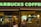 A Starbucks coffee shop with bright green signage. Inside, a man is sitting at a table, holding a cup. The atmosphere inside the shop is warm with soft lighting from hanging lamps. The shop interior includes various coffee-related decor and equipment.