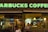 A Starbucks coffee shop with bright green signage. Inside, a man is sitting at a table, holding a cup. The atmosphere inside the shop is warm with soft lighting from hanging lamps. The shop interior includes various coffee-related decor and equipment.