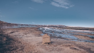 A rocky landscape with a prominent rock labeled 'GEYSIR' in the foreground. Steam rises from the ground, suggesting geothermal activity. The terrain is rugged with patches of snow and water among the rocks, under a clear blue sky.