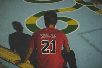 A person wearing a red sports jersey with the name 'Butler' and the number 21 on it is sitting on a wooden surface, viewed from the back. The background features a blue ground with abstract green and yellow patterns, casting a shadow of the person.