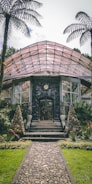 A glass greenhouse with an arched roof stands surrounded by lush green plants and trees. Tall stone cones flank the entrance, which is accessed by a stone pathway. The structure is made of glass and metal, with several windows visible. Two large palm trees frame the scene, adding to the tropical feel.