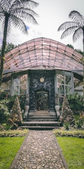 A glass greenhouse with an arched roof stands surrounded by lush green plants and trees. Tall stone cones flank the entrance, which is accessed by a stone pathway. The structure is made of glass and metal, with several windows visible. Two large palm trees frame the scene, adding to the tropical feel.