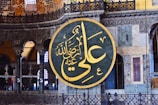A large circular Arabic calligraphy plaque with intricate golden Arabic script hangs prominently on an ornate interior wall. The background showcases detailed architectural features, including decorative arches, columns, and stained glass windows in various shades of blue and yellow.