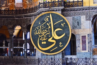A large circular Arabic calligraphy plaque with intricate golden Arabic script hangs prominently on an ornate interior wall. The background showcases detailed architectural features, including decorative arches, columns, and stained glass windows in various shades of blue and yellow.