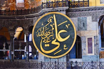 A large circular Arabic calligraphy plaque with intricate golden Arabic script hangs prominently on an ornate interior wall. The background showcases detailed architectural features, including decorative arches, columns, and stained glass windows in various shades of blue and yellow.
