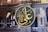 A large circular Arabic calligraphy plaque with intricate golden Arabic script hangs prominently on an ornate interior wall. The background showcases detailed architectural features, including decorative arches, columns, and stained glass windows in various shades of blue and yellow.