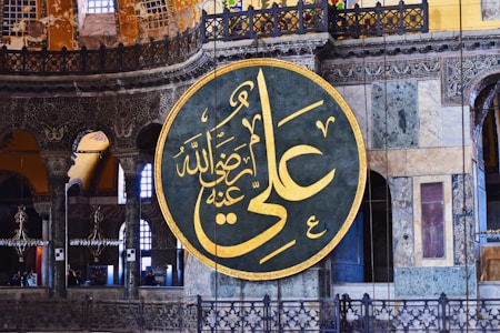 A large circular Arabic calligraphy plaque with intricate golden Arabic script hangs prominently on an ornate interior wall. The background showcases detailed architectural features, including decorative arches, columns, and stained glass windows in various shades of blue and yellow.