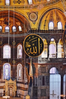 An ornate interior with large circular Islamic calligraphy panels featuring Arabic script. The architecture includes arched windows, elaborate patterns, and intricate details on the walls, with a yellow and brown color scheme dominating the walls and ceiling.