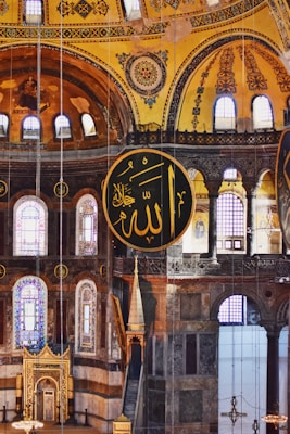 An ornate interior with large circular Islamic calligraphy panels featuring Arabic script. The architecture includes arched windows, elaborate patterns, and intricate details on the walls, with a yellow and brown color scheme dominating the walls and ceiling.