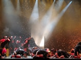 A group of performers on stage, dressed in colorful costumes, appear to be engaged in a dramatic or energetic dance routine. The background is illuminated with bright spotlights and speckled with what looks like falling confetti or theatrical effects, adding to the dynamic atmosphere of the scene.