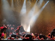A group of performers on stage, dressed in colorful costumes, appear to be engaged in a dramatic or energetic dance routine. The background is illuminated with bright spotlights and speckled with what looks like falling confetti or theatrical effects, adding to the dynamic atmosphere of the scene.