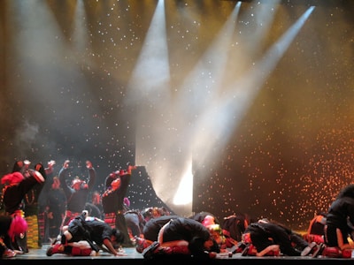 A group of performers on stage, dressed in colorful costumes, appear to be engaged in a dramatic or energetic dance routine. The background is illuminated with bright spotlights and speckled with what looks like falling confetti or theatrical effects, adding to the dynamic atmosphere of the scene.