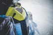 A snowmobile with a yellow hood is moving through snow, creating a spray of snow particles in the air. The surface of the snow is smooth, and the snowmobile's design details, such as the vent and cautionary label, are visible.