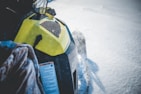 A snowmobile with a yellow hood is moving through snow, creating a spray of snow particles in the air. The surface of the snow is smooth, and the snowmobile's design details, such as the vent and cautionary label, are visible.