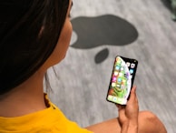 A person wearing a yellow shirt is holding a smartphone in their hand, displaying colorful app icons on the screen. The background features a grey floor with a large black silhouette of a stylized apple.