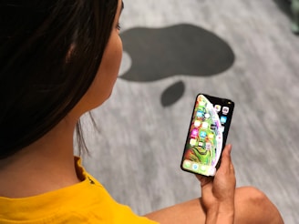 A person wearing a yellow shirt is holding a smartphone in their hand, displaying colorful app icons on the screen. The background features a grey floor with a large black silhouette of a stylized apple.