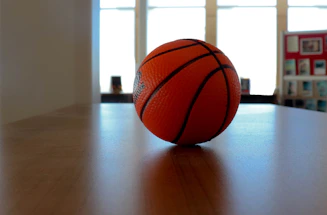 A close-up of a basketball resting on a wooden gym floor with a blurred flyer in the background.