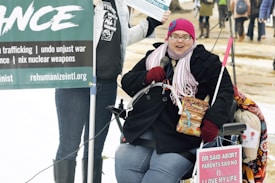 A person wearing a pink knitted hat, glasses, and a scarf is seated in a wheelchair. They are holding a microphone and a box of Cracker Jacks. There are protest signs around them, including one that highlights anti-violence and anti-war messages. The background shows a mix of people standing and walking in an outdoor setting with some snow on the ground.