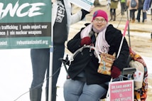 A person wearing a pink knitted hat, glasses, and a scarf is seated in a wheelchair. They are holding a microphone and a box of Cracker Jacks. There are protest signs around them, including one that highlights anti-violence and anti-war messages. The background shows a mix of people standing and walking in an outdoor setting with some snow on the ground.