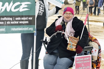 A person wearing a pink knitted hat, glasses, and a scarf is seated in a wheelchair. They are holding a microphone and a box of Cracker Jacks. There are protest signs around them, including one that highlights anti-violence and anti-war messages. The background shows a mix of people standing and walking in an outdoor setting with some snow on the ground.