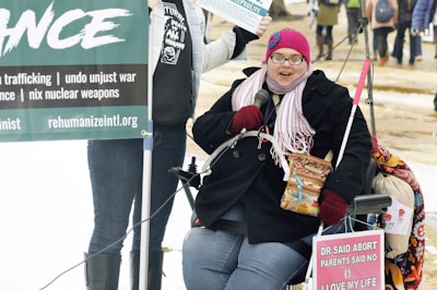A person wearing a pink knitted hat, glasses, and a scarf is seated in a wheelchair. They are holding a microphone and a box of Cracker Jacks. There are protest signs around them, including one that highlights anti-violence and anti-war messages. The background shows a mix of people standing and walking in an outdoor setting with some snow on the ground.