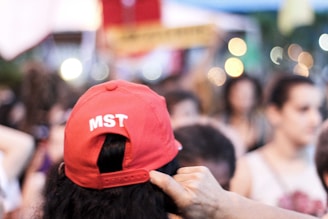 A person is wearing a red cap with 'MST' printed on it, standing amidst a group of people. The background is blurry, suggesting it might be a public gathering or protest. Bright bokeh lights are visible, adding a vibrant touch to the scene.