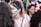 A group of people in a crowd, with a young person having 'Faltam as Mortas' written on their face. The focus is on this individual with pink-tinted hair wearing a choker and a grey top. Other individuals are blurred in the background.