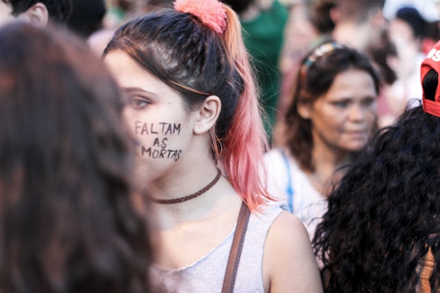 A group of people in a crowd, with a young person having 'Faltam as Mortas' written on their face. The focus is on this individual with pink-tinted hair wearing a choker and a grey top. Other individuals are blurred in the background.