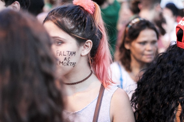 A group of people in a crowd, with a young person having 'Faltam as Mortas' written on their face. The focus is on this individual with pink-tinted hair wearing a choker and a grey top. Other individuals are blurred in the background.