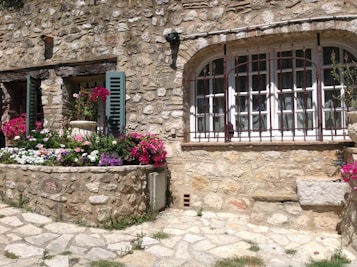 A rustic stone cottage wall with arched windows and wooden shutters, surrounded by vibrant flower beds. The windows are adorned with metal grilles, and the stone path leads to a charming garden area full of colorful blooms.