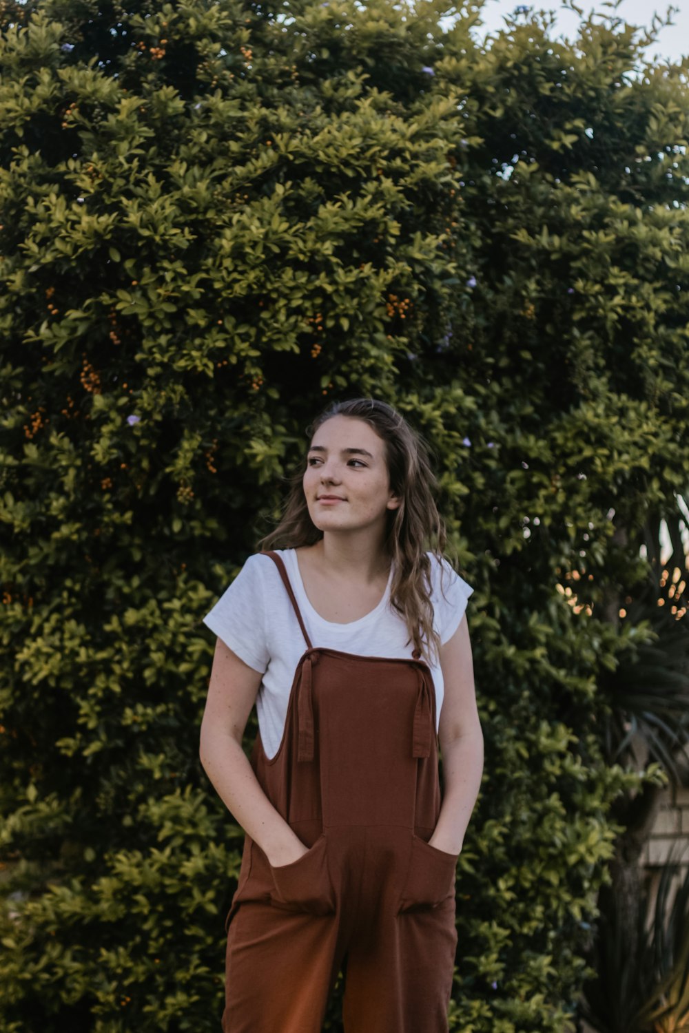 smiling woman wearing brown overall pants standing beside green plant