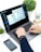 A person is using a MacBook Pro, typing on the keyboard. A smartwatch is visible on their wrist. An iPhone, a pair of AirPods, and a potted plant are placed on the white surface beside the laptop. A take-away coffee cup with the Starbucks logo is in the background.