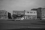 A streetscape view of a commercial area featuring several businesses, including a health center, a shop that buys gold and watches, and a massage therapy center. Cars are parked on the street in front of these buildings. The architecture is a mix of low-rise retail and residential buildings. The scene is captured in black and white, suggesting an overcast day.