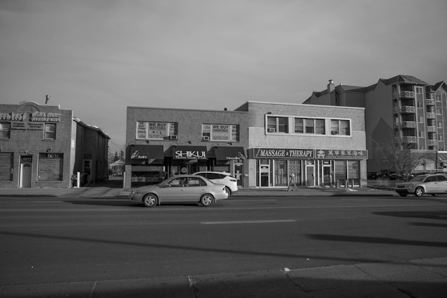 A streetscape view of a commercial area featuring several businesses, including a health center, a shop that buys gold and watches, and a massage therapy center. Cars are parked on the street in front of these buildings. The architecture is a mix of low-rise retail and residential buildings. The scene is captured in black and white, suggesting an overcast day.