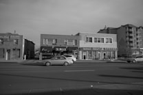 A streetscape view of a commercial area featuring several businesses, including a health center, a shop that buys gold and watches, and a massage therapy center. Cars are parked on the street in front of these buildings. The architecture is a mix of low-rise retail and residential buildings. The scene is captured in black and white, suggesting an overcast day.