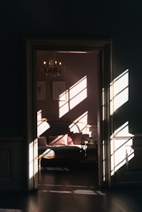 A cozy, elegant room with sunlight streaming through a window, casting patterned shadows on the walls and a luxurious sofa. A chandelier hangs from the ceiling, adding to the sophisticated ambiance, while framed art adorns the walls.