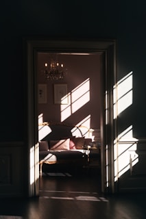 A cozy, elegant room with sunlight streaming through a window, casting patterned shadows on the walls and a luxurious sofa. A chandelier hangs from the ceiling, adding to the sophisticated ambiance, while framed art adorns the walls.