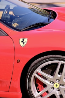 A close-up of a red sports car, focusing on the front wheel and part of the door. The iconic logo is prominently displayed near the wheel. The car's sleek design and vibrant color highlight its luxurious nature.