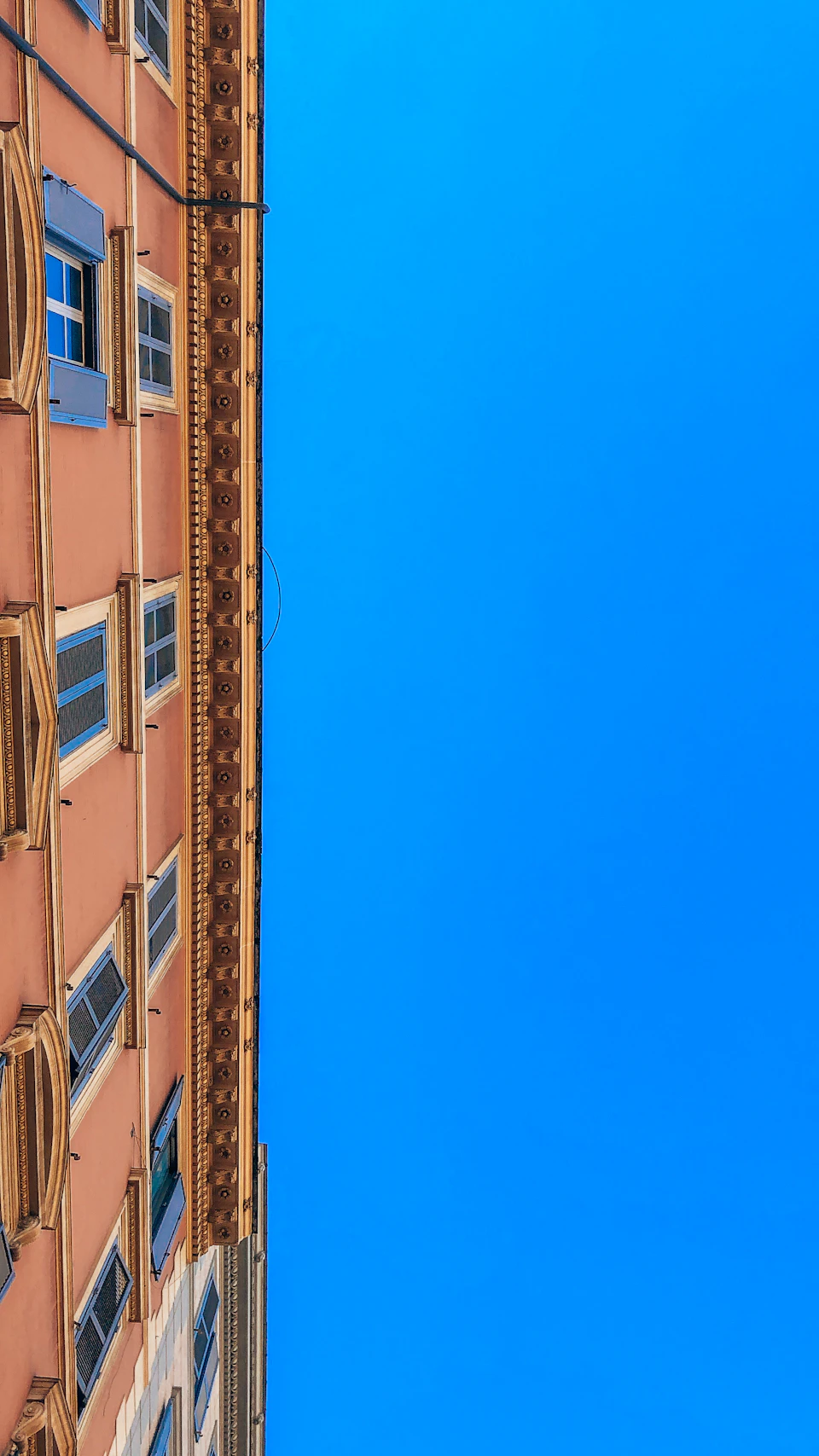 The image shows a tall, narrow building with a pink facade and blue accents on its windows. The perspective is from below, looking up at the structure against a clear blue sky. The building's design features multiple levels, adding to its height and prominence in the scene.