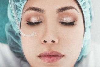 A person with closed eyes is wearing a blue surgical cap. Their eyes are adorned with shimmery makeup, and a transparent tube runs along the side of their face.