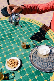 A hand pouring tea from a silver teapot into a glass on a green and yellow tiled table. A plate with various pastries and a decorative tray with sugar cubes are also visible.