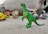A collection of colorful toy dinosaurs is arranged on a surface, with a green dinosaur figurine standing prominently in the foreground. Other dinosaur toys in various colors are slightly blurred in the background, adding depth to the scene.