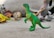 A collection of colorful toy dinosaurs is arranged on a surface, with a green dinosaur figurine standing prominently in the foreground. Other dinosaur toys in various colors are slightly blurred in the background, adding depth to the scene.