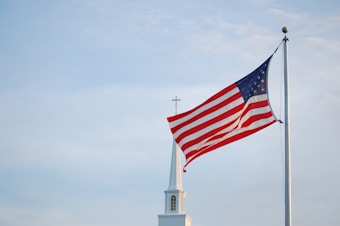 An American flag waves prominently on a tall flagpole against a backdrop of a clear sky. In the background, the steeple of a church is visible, topped with a cross.