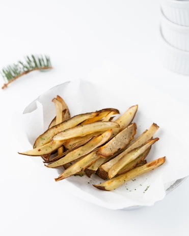 A serving of crispy baked potato wedges garnished with herbs, placed in a white paper-lined basket. The background features a blurred branch element and stacked ceramic dishes.