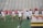 A group of female athletes wearing white tops and yellow skirts are on a lacrosse field. Some are holding lacrosse sticks and running or walking across the field toward a goalpost. The setting appears to be a sports stadium with empty bleachers in the background.