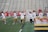A group of female athletes wearing white tops and yellow skirts are on a lacrosse field. Some are holding lacrosse sticks and running or walking across the field toward a goalpost. The setting appears to be a sports stadium with empty bleachers in the background.